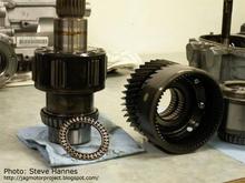 The assembly seen below the main drive gear on the mainshaft is the planetary gear set. This is the assembly that "splits" the incoming torque from the transmission into the 61%/39% ratio. Here is what the gearset looks like:  Look closely and you can see the planetary gears in a carrier on the mainshaft. Next to it on the right is the mating gear housing. Essentially the way this works is: The mainshaft on the left is actually two shafts, one inside another. The outside shaft receives all of the torque from the transmission output.